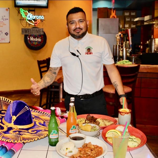 A La Unica staff member stands behind a table laden with Mexican food, drinks, and a sombrero.