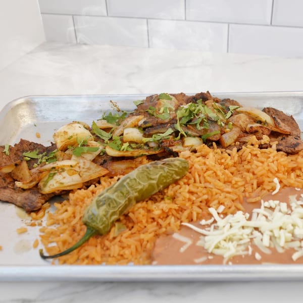 Steaming carne asada with onions, cilantro, rice, beans, cheese, and a jalapeño on a metal tray.