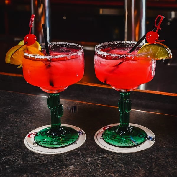 Two vibrant red margaritas in cactus-stemmed glasses, garnished with fruit and cherries, on a bar.