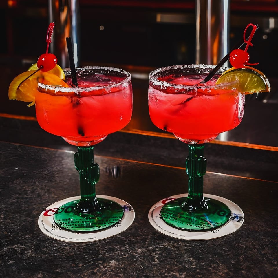 Two vibrant red margaritas in cactus-stemmed glasses, garnished with fruit and cherries, on a bar.