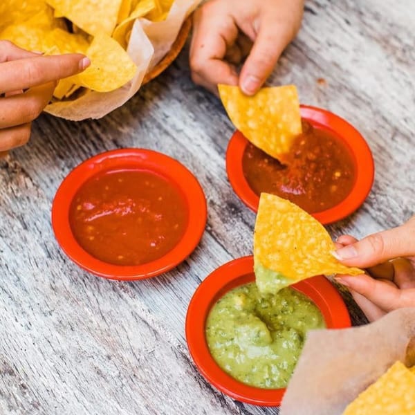 Hands dipping tortilla chips into red salsa and green guacamole on a rustic wooden table.