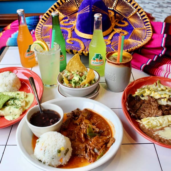 A table spread with La Unica Mexican Restaurant's dishes, Jarritos, and margaritas, with a sombrero.