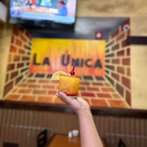 Hand holding a colorful cocktail with a cherry and orange slice, in front of a "La Unica" sign.