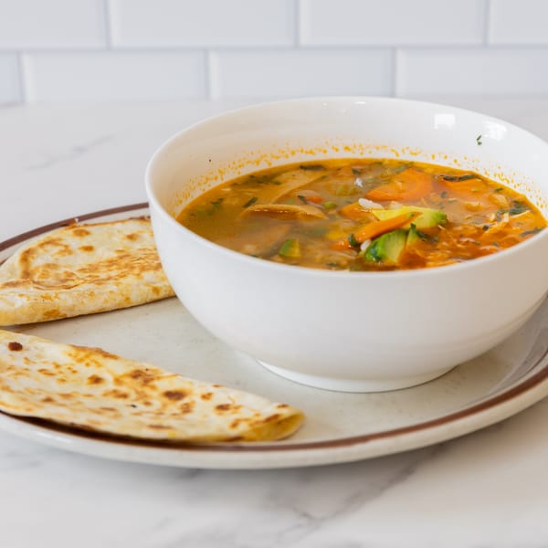 A white bowl of La Unica Mexican Restaurant's vegetable soup with tortillas on a plate.