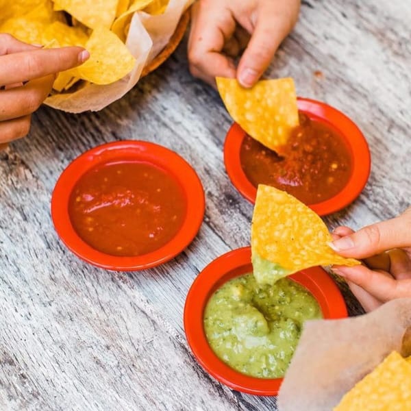 Hands dipping tortilla chips into red salsa and green guacamole in red bowls on a wooden table.