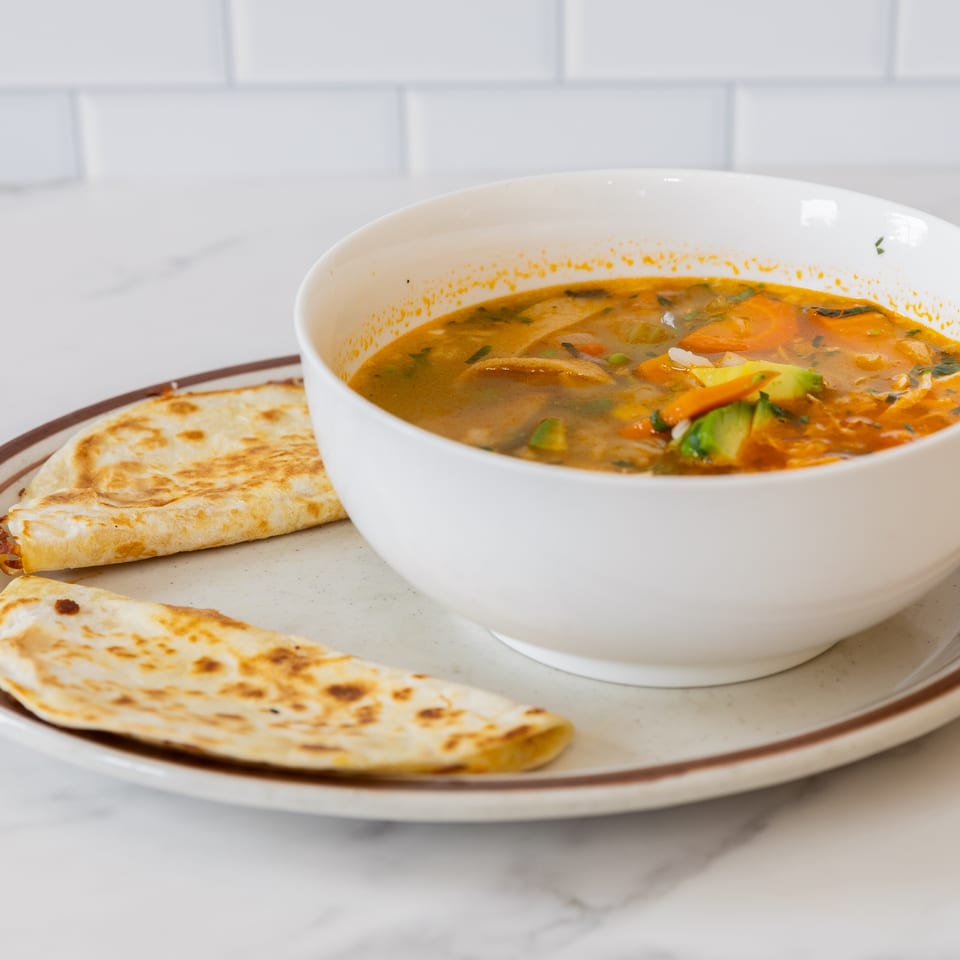 A white bowl of La Unica Mexican Restaurant's vegetable soup with two quesadillas on a plate.
