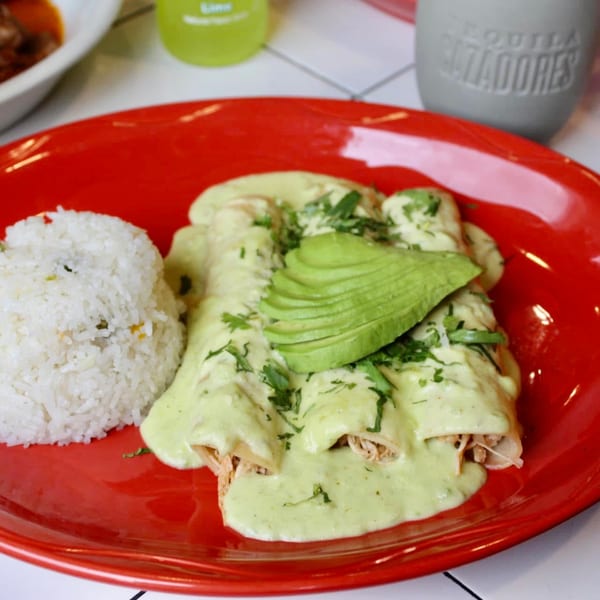 Three chicken enchiladas with green sauce, avocado slices, and a side of white rice on a red plate.
