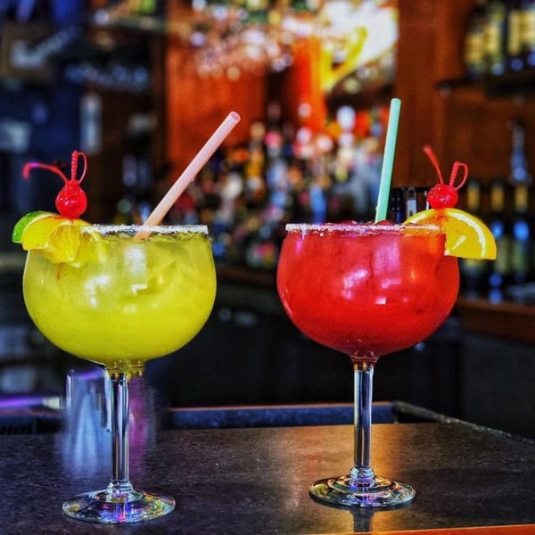 Two vibrant margaritas, one yellow and one red, garnished with fruit and straws on a bar.
